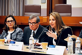 Secretary Hughs sitting at a table with other officials. She is speaking into a microphone and is addressing the room. 